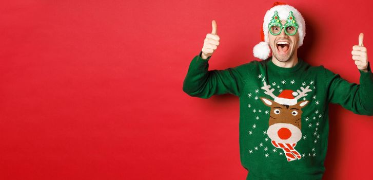 Portrait of super happy young man in party glasses, santa hat and sweater, showing thumbs-up in approval, recommending new year promo offer, standing over red background.