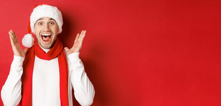 Concept of christmas, winter holidays and celebration. Image of handsome man looking surprised at new year promo offer, smiling amazed, standing over red background.