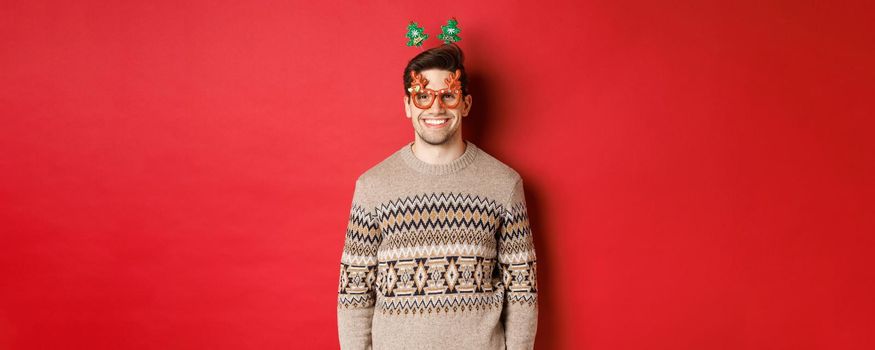Portrait of handsome bearded guy in party glasses and winter sweater, smiling pleased, celebrating new year, wishing merry christmas, standing over red background.