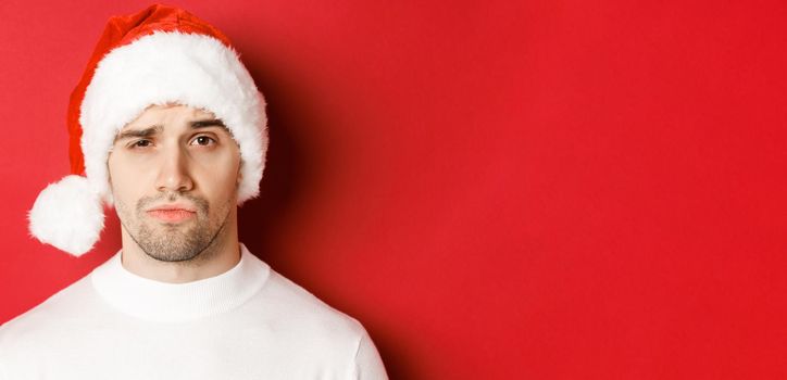 Close-up of doubtful frowning man, wearing santa hat and looking at camera uncertain, standing against red background.