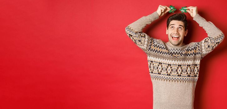 Concept of winter holidays, christmas and celebration. Image of funny and cute adult man enjoying new year party, smiling and rejoicing, standing over red background.