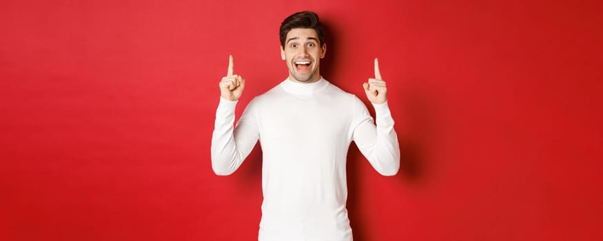 Concept of winter holidays. Good-looking young man with bristle, wearing white sweater, showing christmas advertisement on copy space, pointing fingers up and looking amazed, red background.