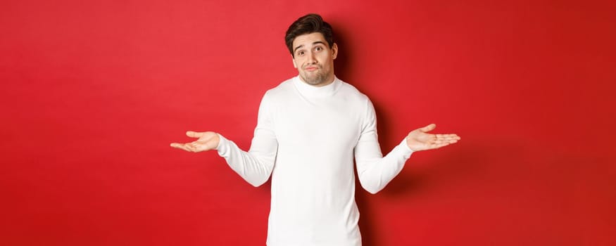 Portrait of clueless handsome guy, shrugging with hands spread sideways, being unaware, dont know anything, standing over red background.