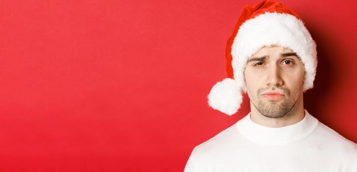 Close-up of doubtful frowning man, wearing santa hat and looking at camera uncertain, standing against red background.