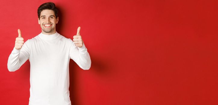 Concept of winter holidays, christmas and lifestyle. Portrait of satisfied handsome man in white sweater showing thumbs-up, praise something good, like it, standing over red background.