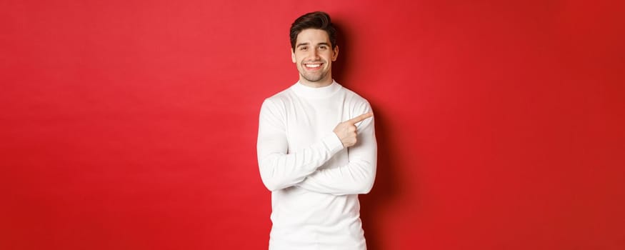Handsome young man with bristle, wearing white sweater, pointing fingers at upper right corner and smiling, showing christmas banner, standing over red background.