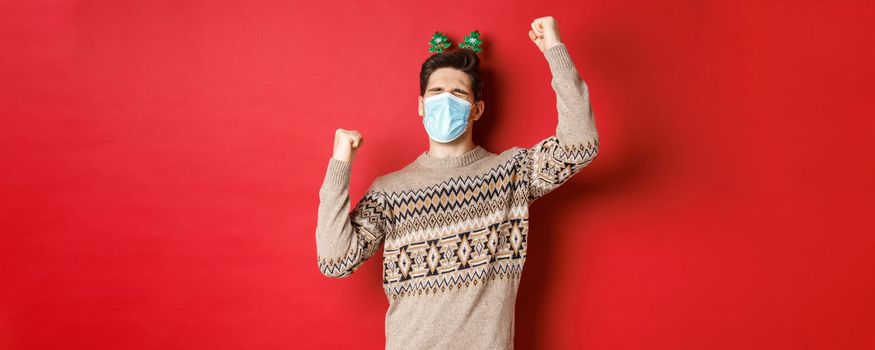 Concept of coronavirus, winter holidays and social distancing. Image of excited and happy man in medical mask and christmas clothing, raising hands up and celebrating new year, red background.