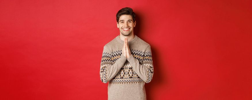 Image of happy and cute man in christmas sweater, asking for favour, holding hands together, smiling and saying thank you, feeling grateful, standing over red background.