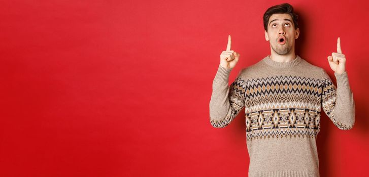 Image of amazed handsome man in christmas sweater, pointing and looking at new year promo offer, showing winter holidays advertisement on red copy space.