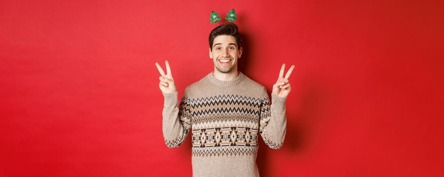 Concept of winter holidays, christmas and celebration. Image of handsome and silly guy dressed for new year party, showing peace signs and smiling, standing against red background.
