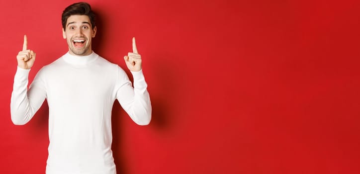 Concept of winter holidays. Good-looking young man with bristle, wearing white sweater, showing christmas advertisement on copy space, pointing fingers up and looking amazed, red background.