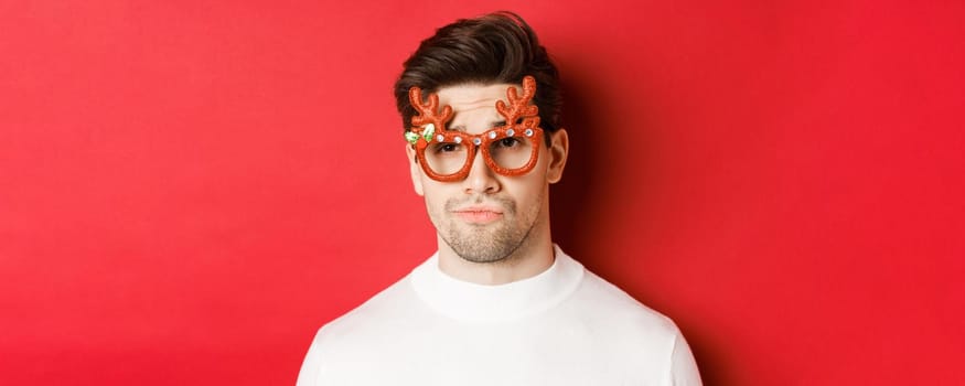 Concept of winter holidays, christmas and celebration. Close-up of skeptical handsome guy, wearing party glasses, smirk and looking unamused, standing over red background.