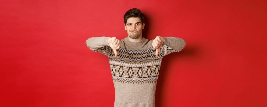Portrait of skeptical and disappointed handsome man in christmas sweater, dislike new year party, showing thumbs-down, express disapproval, standing over red background.