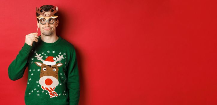 Funny man in christmas sweater and party mask, celebrating winter holidays, showing funny faces, standing against red background.