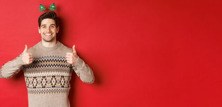 Concept of winter holidays, christmas and celebration. Cheerful bearded guy in sweater, showing thumbs-up in approval and smiling, enjoying new year party, red background.