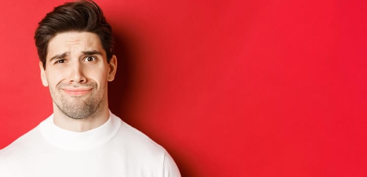 Close-up of handsome man feeling uncomfortable, grimacing and looking at something displeasing, standing over red background.