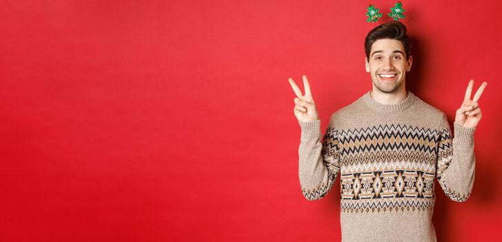 Concept of winter holidays, christmas and celebration. Image of handsome and silly guy dressed for new year party, showing peace signs and smiling, standing against red background.