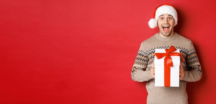Image of excited handsome man receiving christmas gift, wearing santa hat and winter sweater, shouting for joy, holding present and standing over red background.