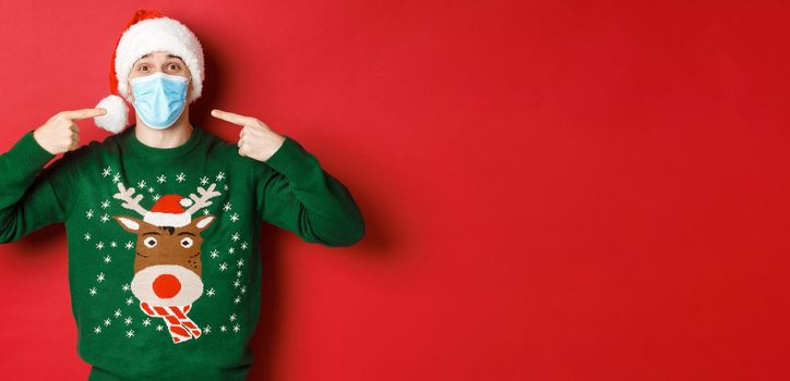 Concept of new year, coronavirus and social distancing. Happy man in santa hat and christmas sweater, recommending to wear medical mask on party, standing over red background.