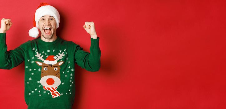 Portrait of cheerful attractive man celebrating new year, wearing green sweater and santa hat, shouting for joy, winning or triumphing, standing over red background.