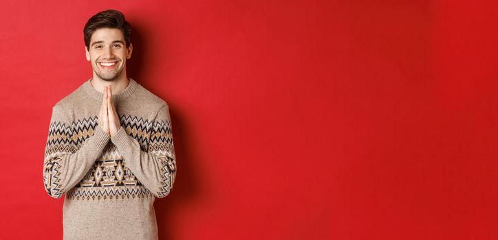 Image of happy and cute man in christmas sweater, asking for favour, holding hands together, smiling and saying thank you, feeling grateful, standing over red background.