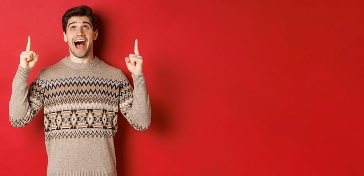 Image of excited handsome guy who likes winter holidays showing advertisement, looking and pointing fingers up with amazed face, standing in christmas sweater over red background.