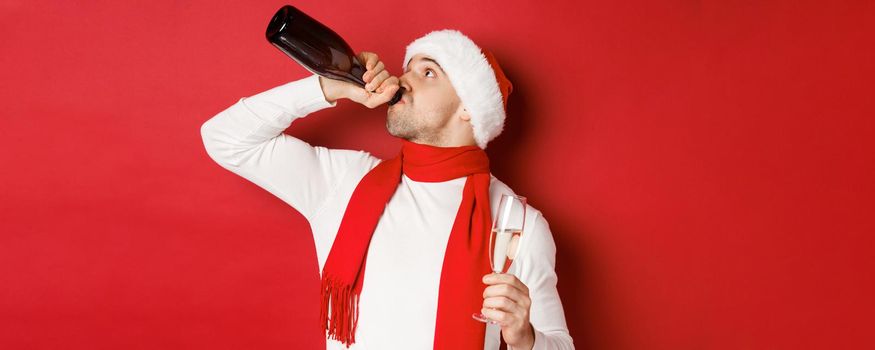 Concept of winter holidays, christmas and lifestyle. Man getting drunk on new year party, drinking champagne from bottle, wearing santa hat, standing over red background.