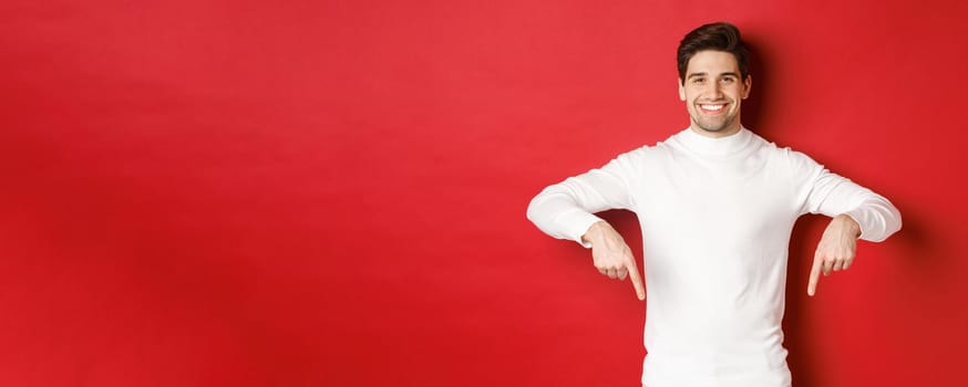 Image of smiling handsome man in white sweater inviting visit page, pointing fingers down and showing christmas banner, standing against red background.