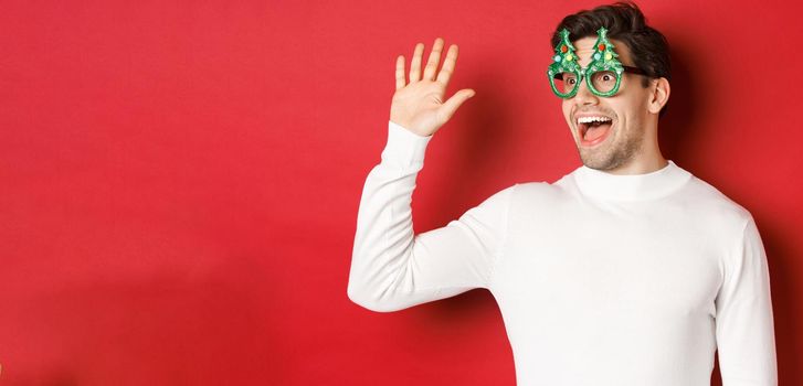 Portrait of friendly, cheerful man in party glasses and white sweater, saying hello and looking left, greeting a friend, standing over red background.