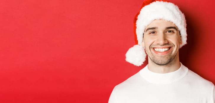 Close-up of attractive smiling man in white sweater and santa hat, looking happy, enjoying winter holidays, standing against red background.