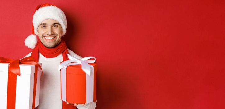 Concept of winter holidays, christmas and lifestyle. Close-up of smiling handsome guy in santa hat and scarf, wishing happy new year and holding gifts, standing with presents over red background.