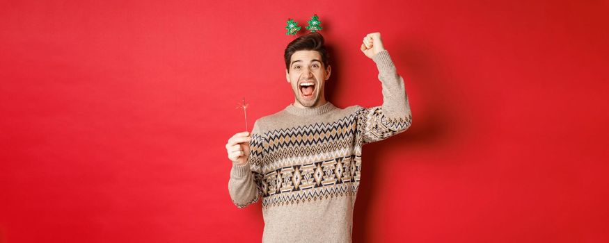 Cheerful guy with a sparkler rejoicing and wishing happy new year, raising hand up and shouting for joy, standing over red background.