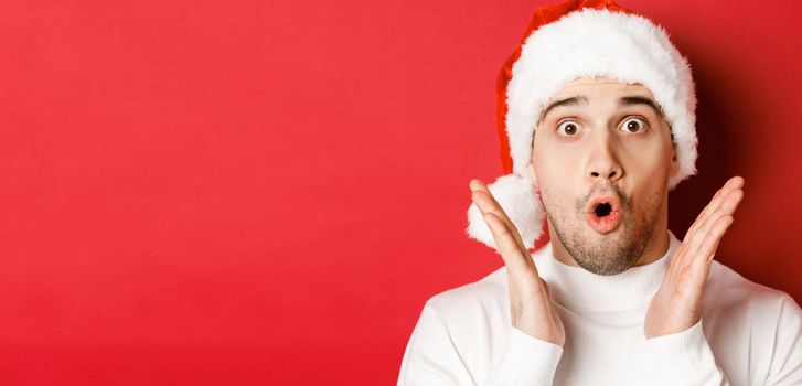 Close-up of amazed handsome man in santa hat and white sweater, raising hands and looking impressed, standing over red background.