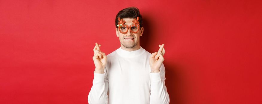 Close-up of handsome nervous guy in party glasses, making a wish, crossing fingers for good luck and looking with hope at camera, standing over red background.