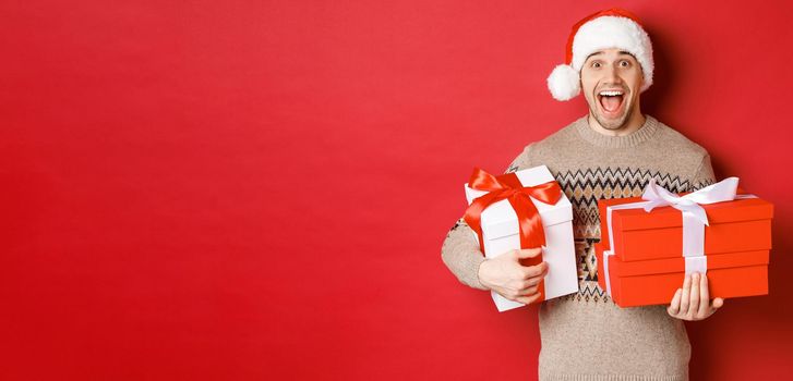 Concept of winter holidays, new year and celebration. Image of amazed and happy man in sweater and santa hat, holding christmas gifts and shouting for joy, red background.