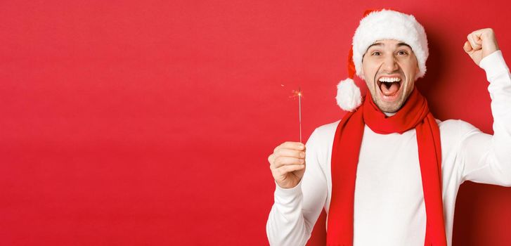 Concept of christmas, winter holidays and celebration. Portrait of excited handsome man, raising hand up and holding sparkler, wishing happy new year, standing over red background.