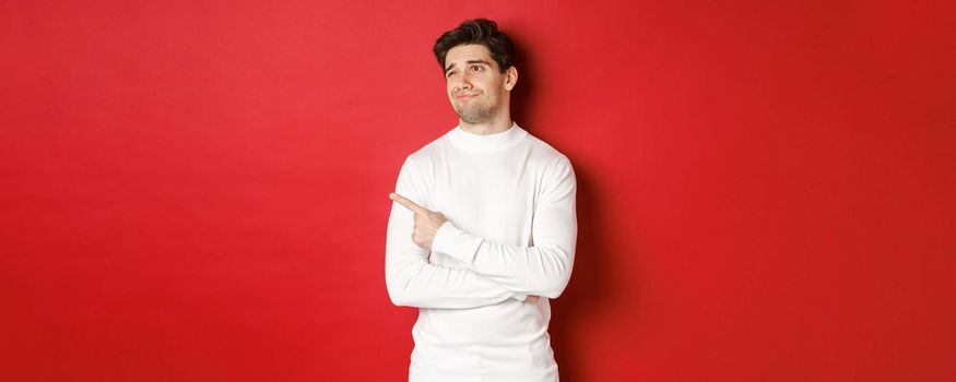 Concept of winter holidays. Image of displeased young man in white sweater, looking and pointing finger left at something average, standing skeptical over red background.