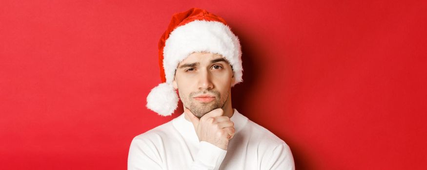 Close-up of thoughtful handsome man in santa hat, frowning and looking at camera, thinking about something, standing over red background.