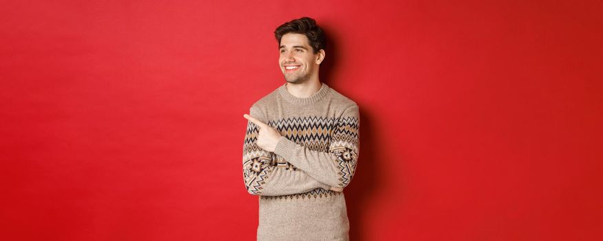 Portrait of attractive caucasian man celebrating new year, winter holidays, pointing finger and looking left with pleased smile, showing christmas advertisement, standing over red background.