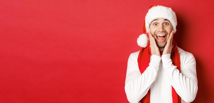 Concept of christmas, winter holidays and celebration. Close-up of surprised and happy man in santa hat and scarf, looking at something amazing, standing over red background.