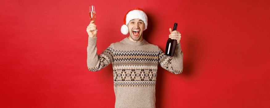 Concept of winter holidays, christmas and celebration. Excited handsome guy in sweater and santa hat, enjoying new year party, raising glass and champagne bottle, having fun.