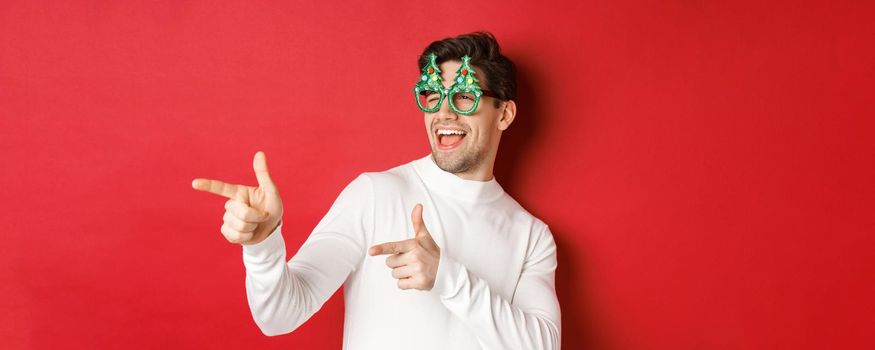 Close-up of cheeky handsome man in party glasses, pointing finger left and greeting person, wishing merry christmas, standing over red background.