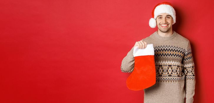 Concept of winter holidays, new year and celebration. Image of handsome smiling man in santa hat and sweater, holding christmas stocking for presents and candies, standing over red background.