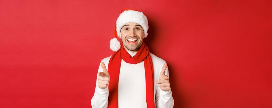 Concept of christmas, winter holidays and celebration. Cheeky man in santa hat and scarf, smiling and pointing fingers at camera, wishing happy new year, standing over red background.