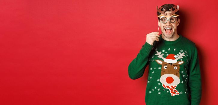 Handsome surprised man in christmas sweater, holding party mask and smiling, enjoying new year celebration, standing over red background.