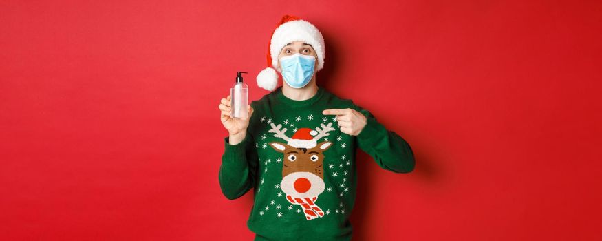 Concept of new year, coronavirus and social distancing. Portrait of cheerful man in santa hat, christmas sweater and medical mask, pointing finger at hand sanitizer, standing over red background.