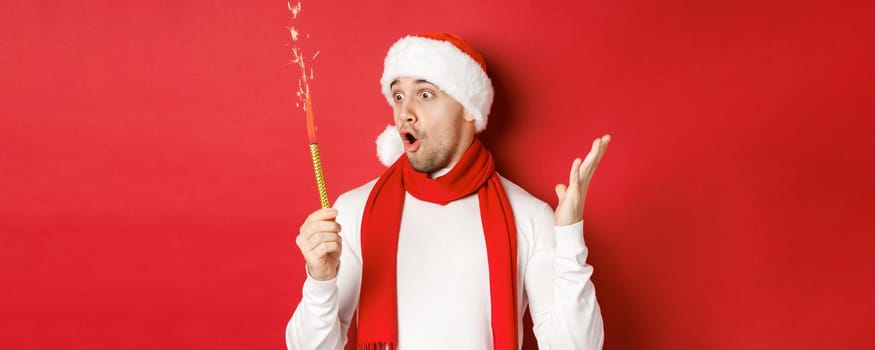 Concept of christmas, winter holidays and celebration. Close-up of amazed handsome man in santa hat and scarf, looking at sparkler with excitement, standing over red background.