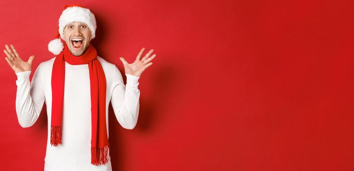 Portrait of happy and amazed handsome man, celebrating new year, wishing merry christmas, wearing santa hat and scarf, telling big news, standing against red background.