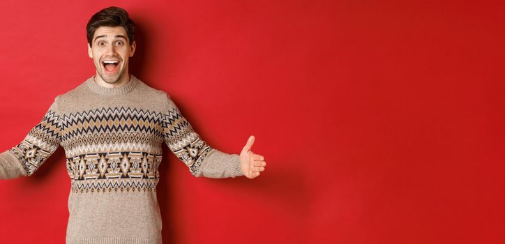 Portrait of attractive happy man in christmas sweater showing big present, spread hands sideways as if holding large box, standing over red background.