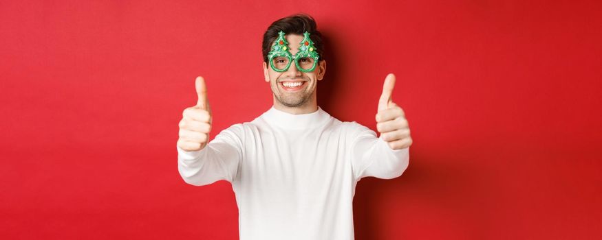 Close-up of attractive happy man in party glasses and white sweater, showing thumbs-up in approval and smiling, standing over red background.